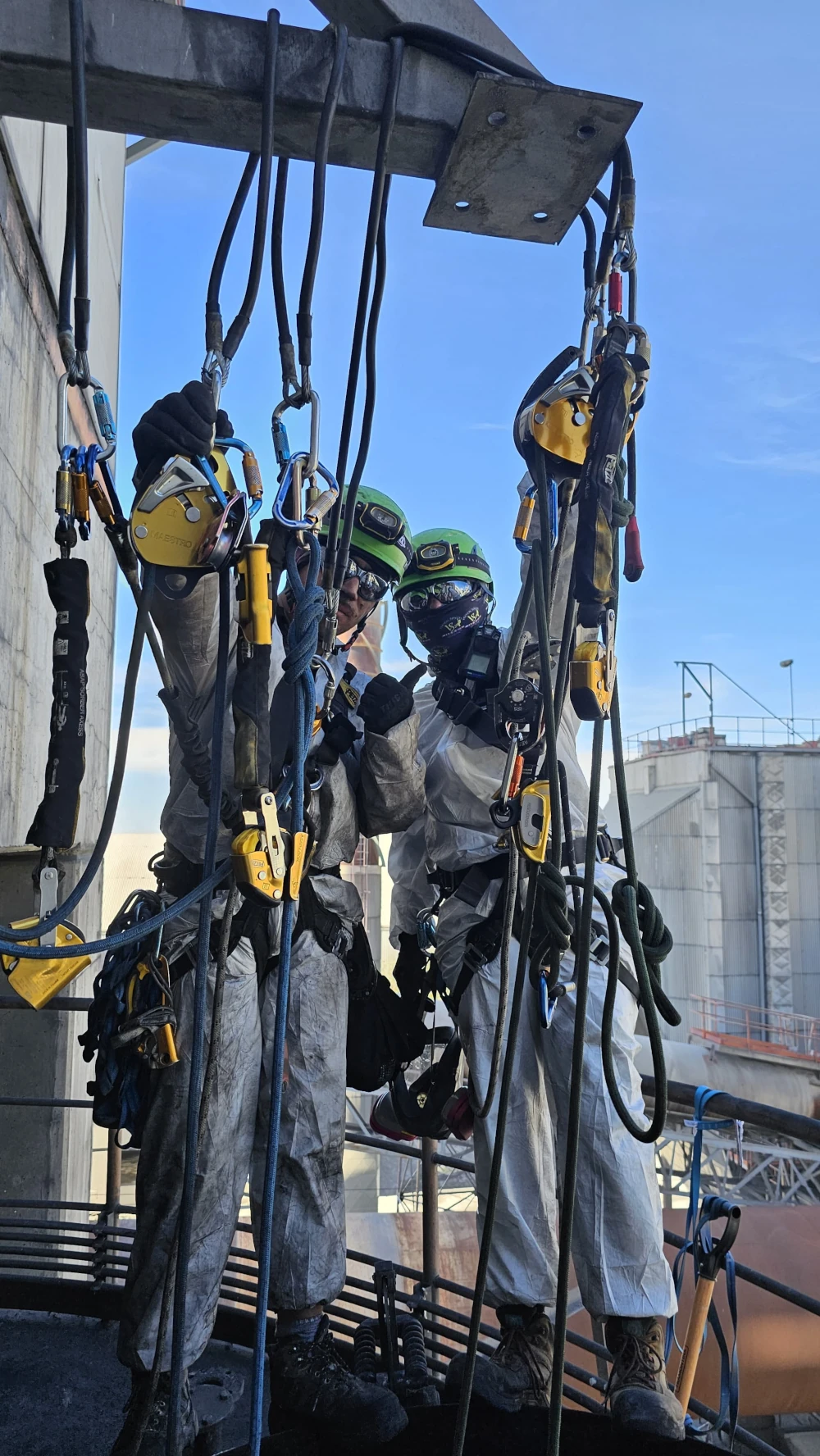 Dos trabajadores industriales de Vertical Service con equipos de protección completos, incluyendo cascos y trajes protectores, suspendidos en el aire con arneses y cuerdas en una estructura industrial.